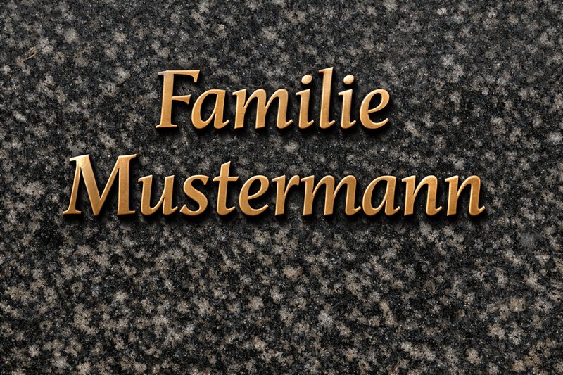 Bronzeschriften auf einem Granitgrabstein: erhabene Buchstaben 'Familie Mustermann' mit warmer Patina, als Beispiel für Metallschriften