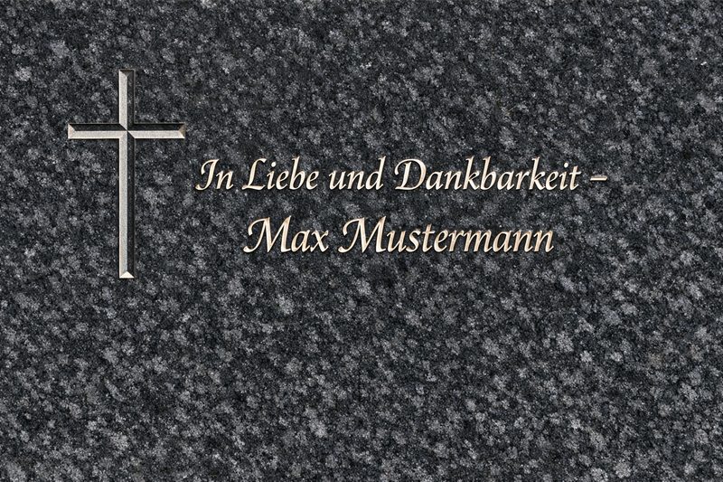 Impala‑Granit mit Keilnut‑Schriftzug 'In Liebe und Dankbarkeit – Max Mustermann' und Kreuz – harmonisch angeordnet im dunklen Naturstein